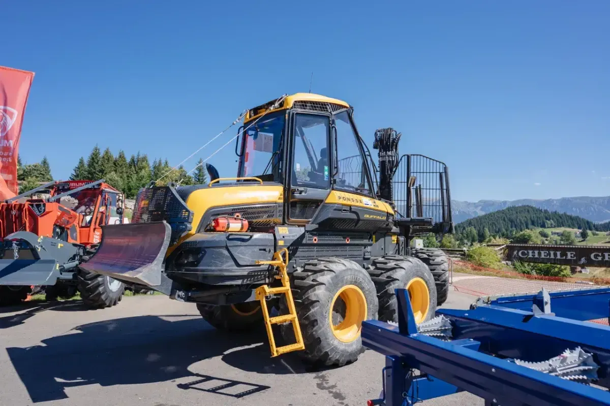 Tractor forestier modern, galben, expus la un târg în aer liber, cu munții în fundal