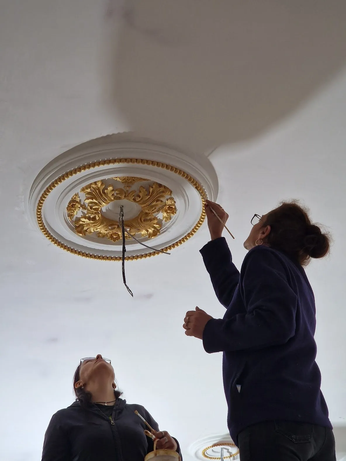 Restauratoare pe schelă aurind cu pensula o rozetă de tavan, în timp ce o colegă privește de jos