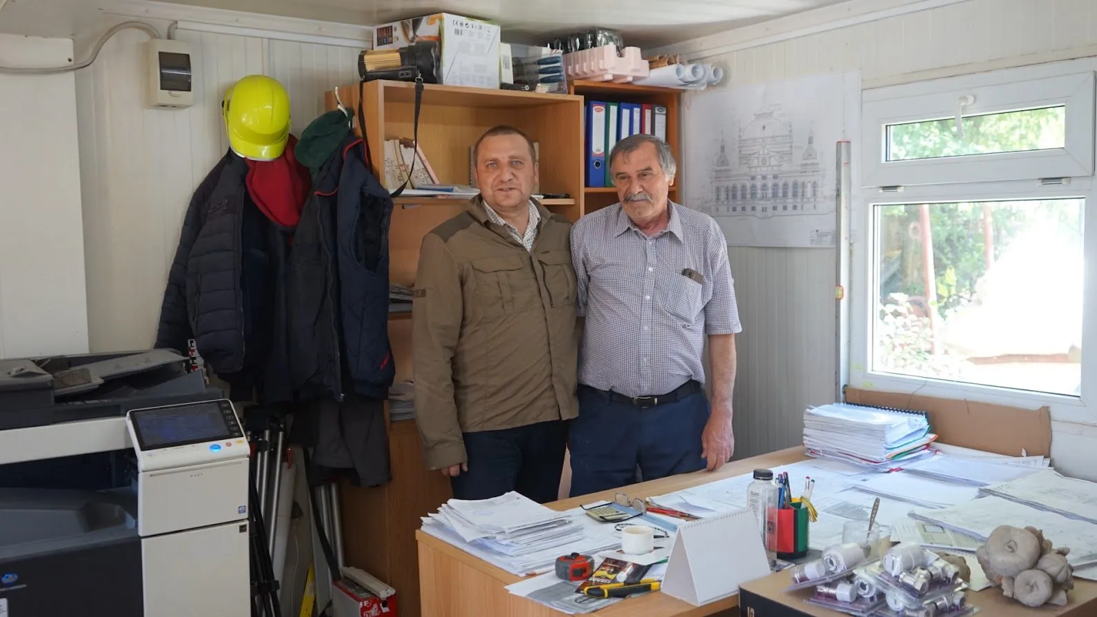 Doi bărbați într-un birou de șantier, cu dosare, planșe și o cască galbenă pe perete; în spatele lor, desenul fațadei Cazinoului