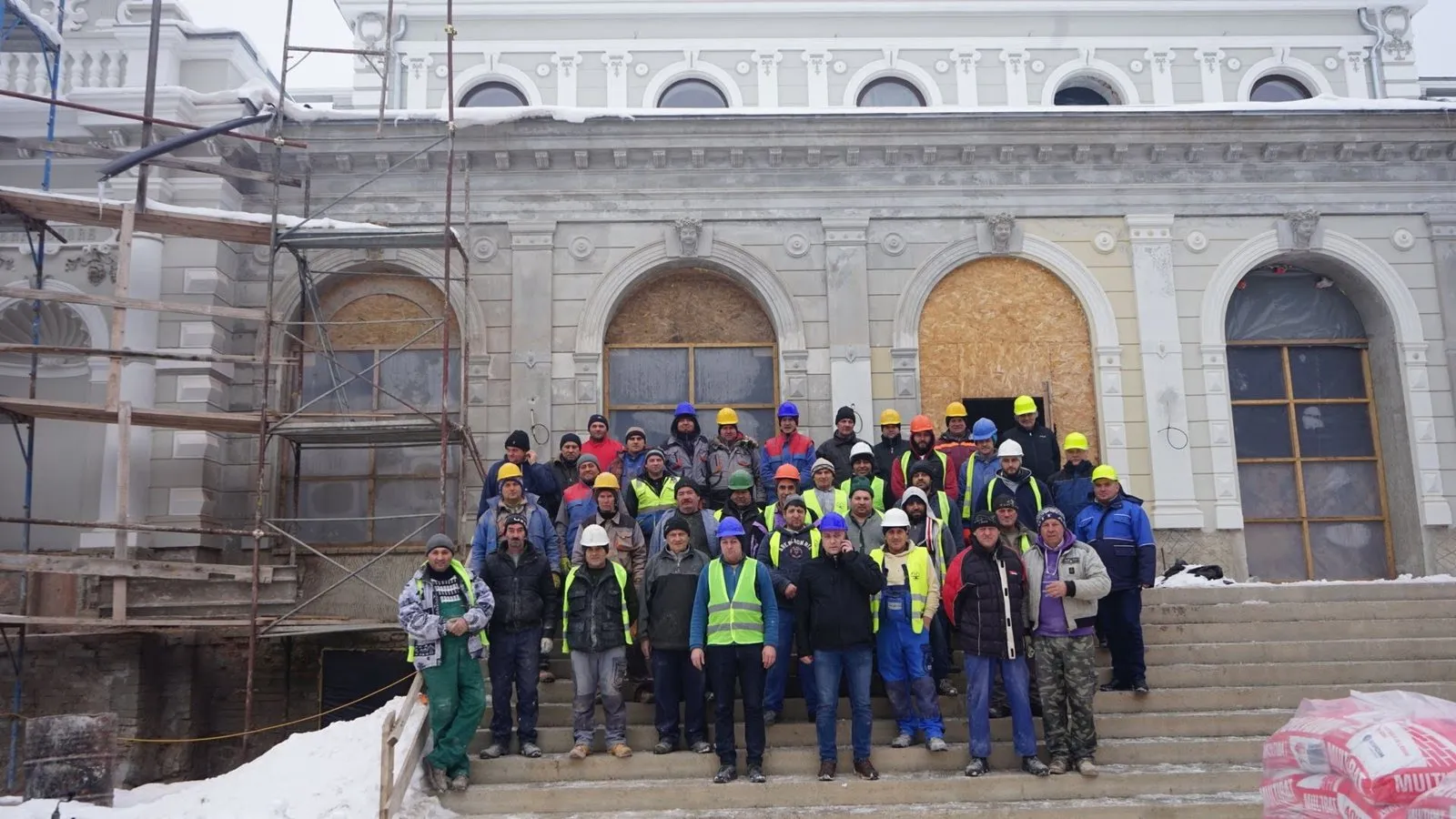 Câteva zeci de muncitori în echipament de lucru, cu căști și veste reflectorizante, pe treptele Cazinoului, iarna, cu zăpadă pe margini