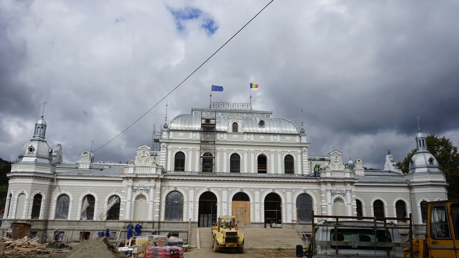 Fațada principală a Cazinoului aproape terminată, sub cer înnorat, cu un utilaj galben în fața scărilor și o schelă pe corpul central