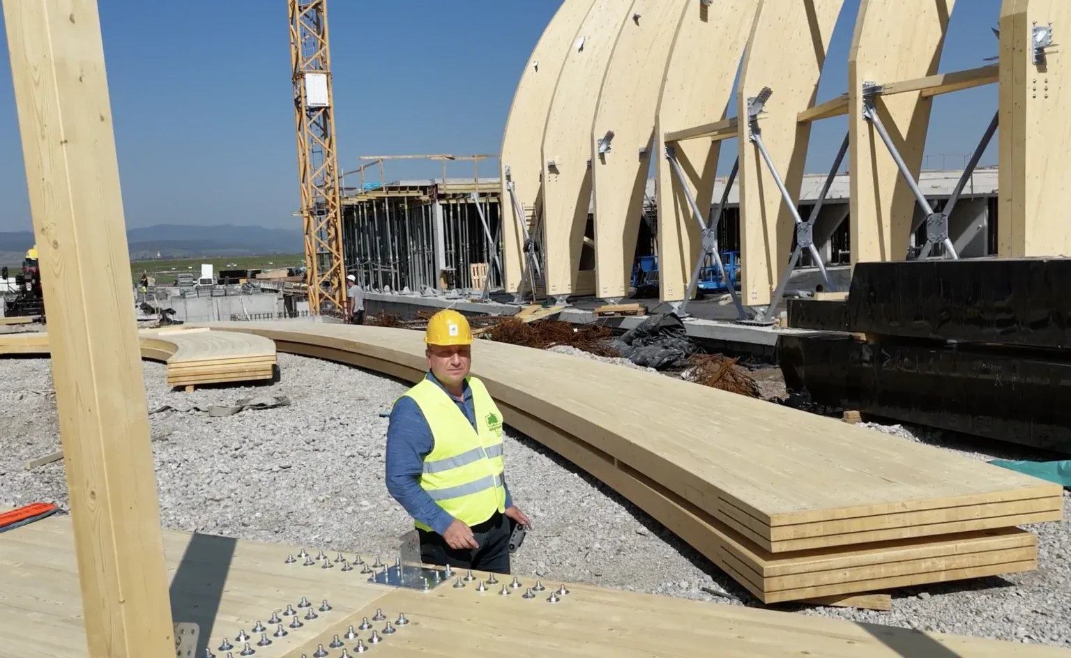 Șantier cu grinzi curbe din lemn lamelat încleiat (glulam) și un muncitor cu cască și vestă reflectorizantă