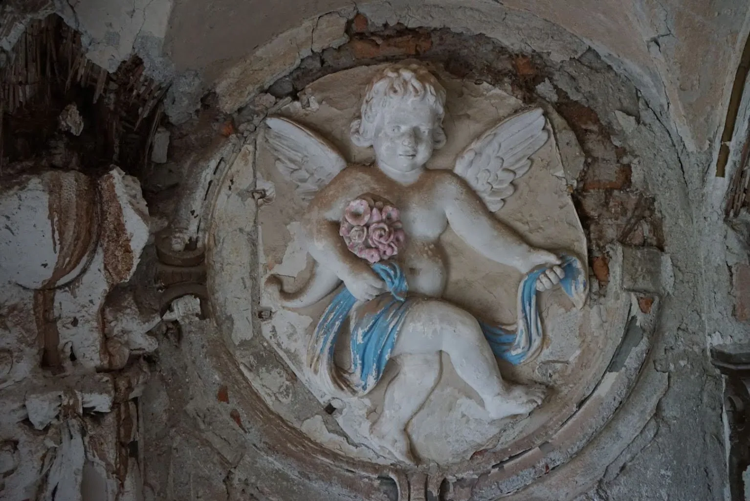 Medalion de stuc cu un înger cu aripi, cu un buchet de trandafiri roz și o eșarfă albastră, înconjurat de tencuială crăpată și cărămidă la vedere
