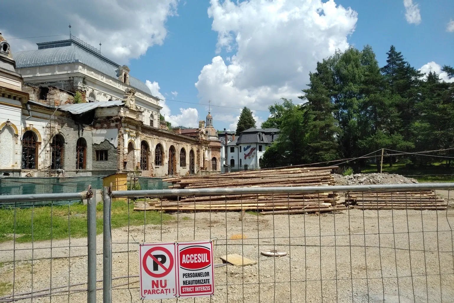Cazinoul Băilor în ruină, văzut de după gardul de șantier, cu stive de bușteni în prim-plan și indicatorul „Accesul persoanelor neautorizate strict interzis”