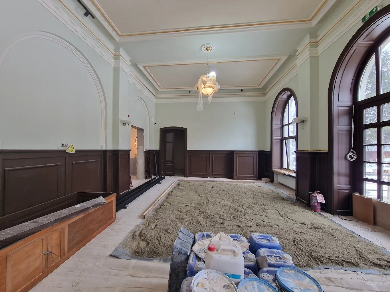 Salon cu lambriuri de lemn închis, ferestre mari în arc și candelabru învelit în folie; pe jos, șapa în lucru și canistre de materiale; afară ninge