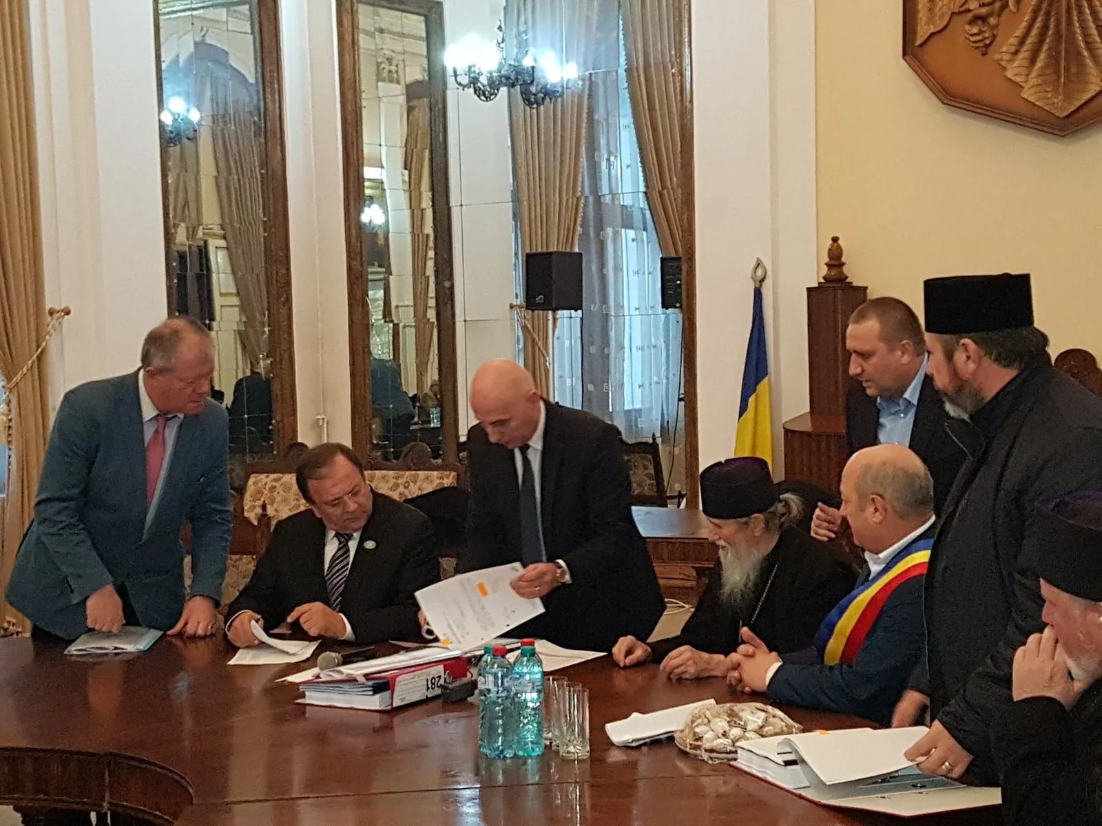 Reprezentanți ai autorităților și ai Arhiepiscopiei semnând documente la o masă lungă, într-o sală cu oglinzi și candelabre