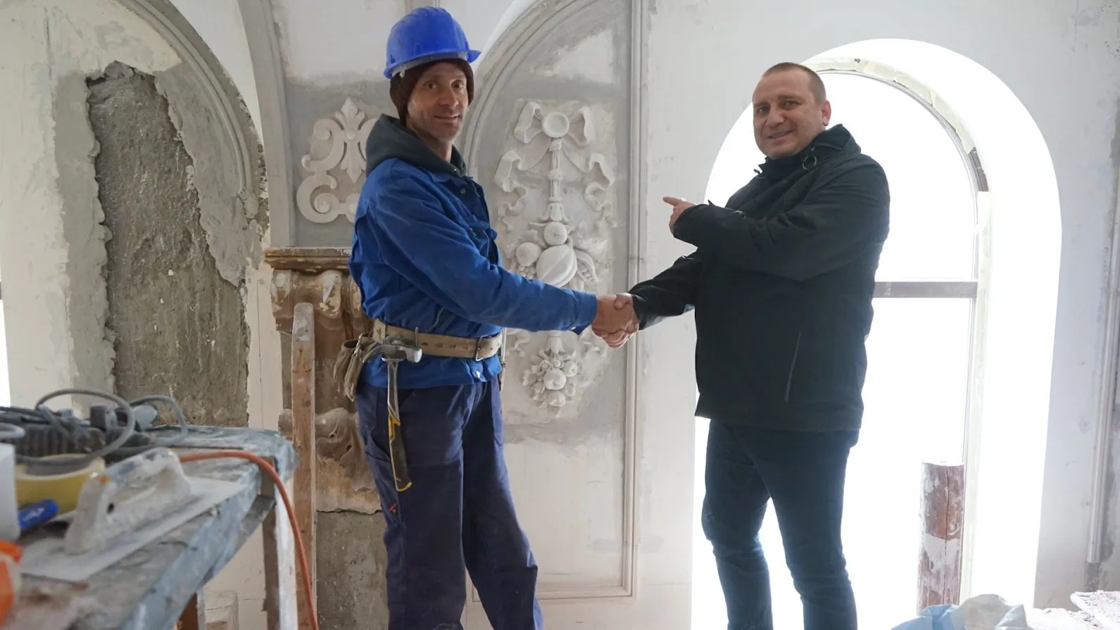 Un stucator cu cască albastră dă mâna cu un bărbat în geacă neagră, care arată spre un ornament de stuc cu panglici și fructe, refăcut pe perete