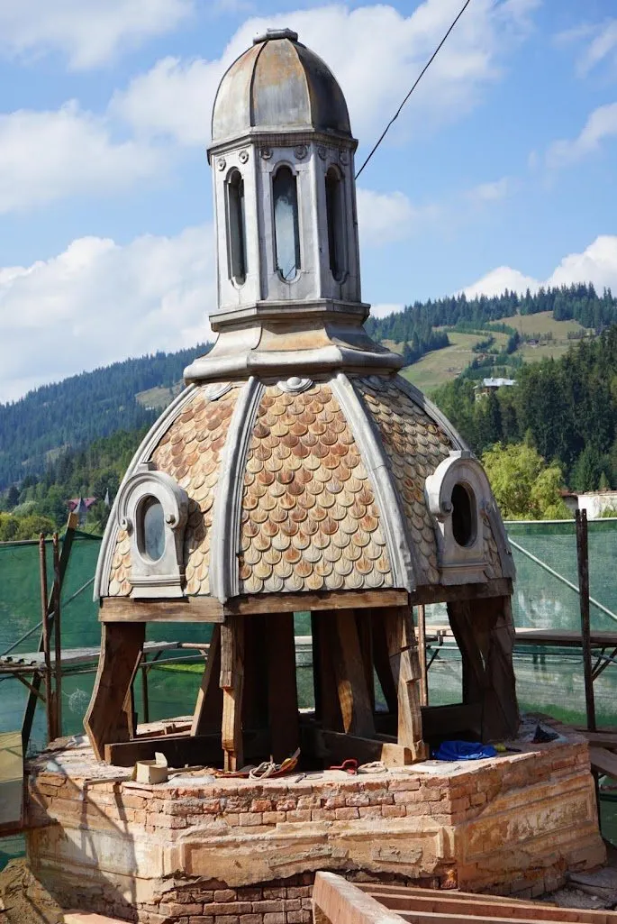 Cupola unui turn de colț în restaurare, cu solzi de tablă și lanternou cu ferestre, așezată pe o bază de cărămidă, cu munții împăduriți în fundal